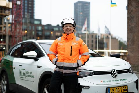 HLM-landmåler i orange arbejdsjakke foran en af HLM's elbiler på en byggeplads i byen